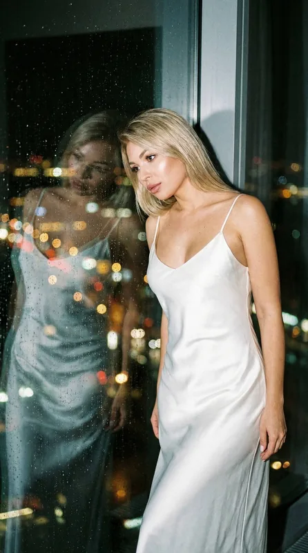 Девушка в длинной белой шолковом приталеном коротком платье на бретелях стоит у большого окна вечером. Огни ночного горо